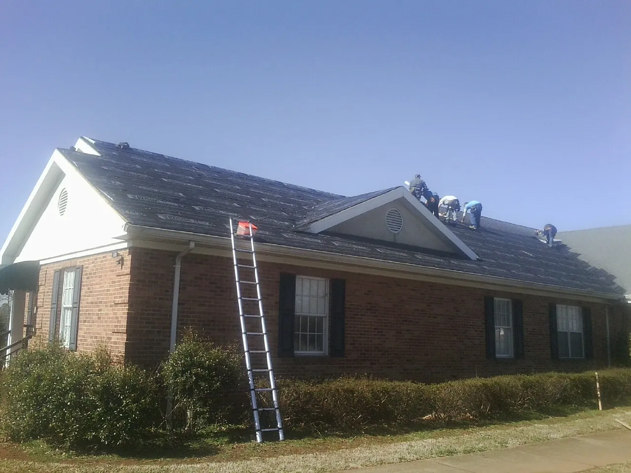 Crew tearing shingles off a brick ranch roof with a ladder set — Emergency Roof Repair work by our crew in Amma