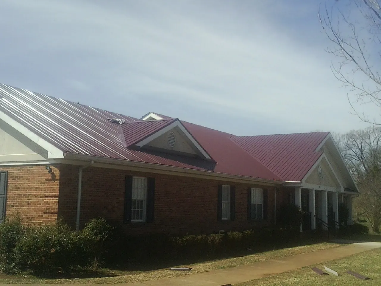 Red metal roof with gabled entries on a long brick building, from a Hail Damage Roof Repair job in Amma