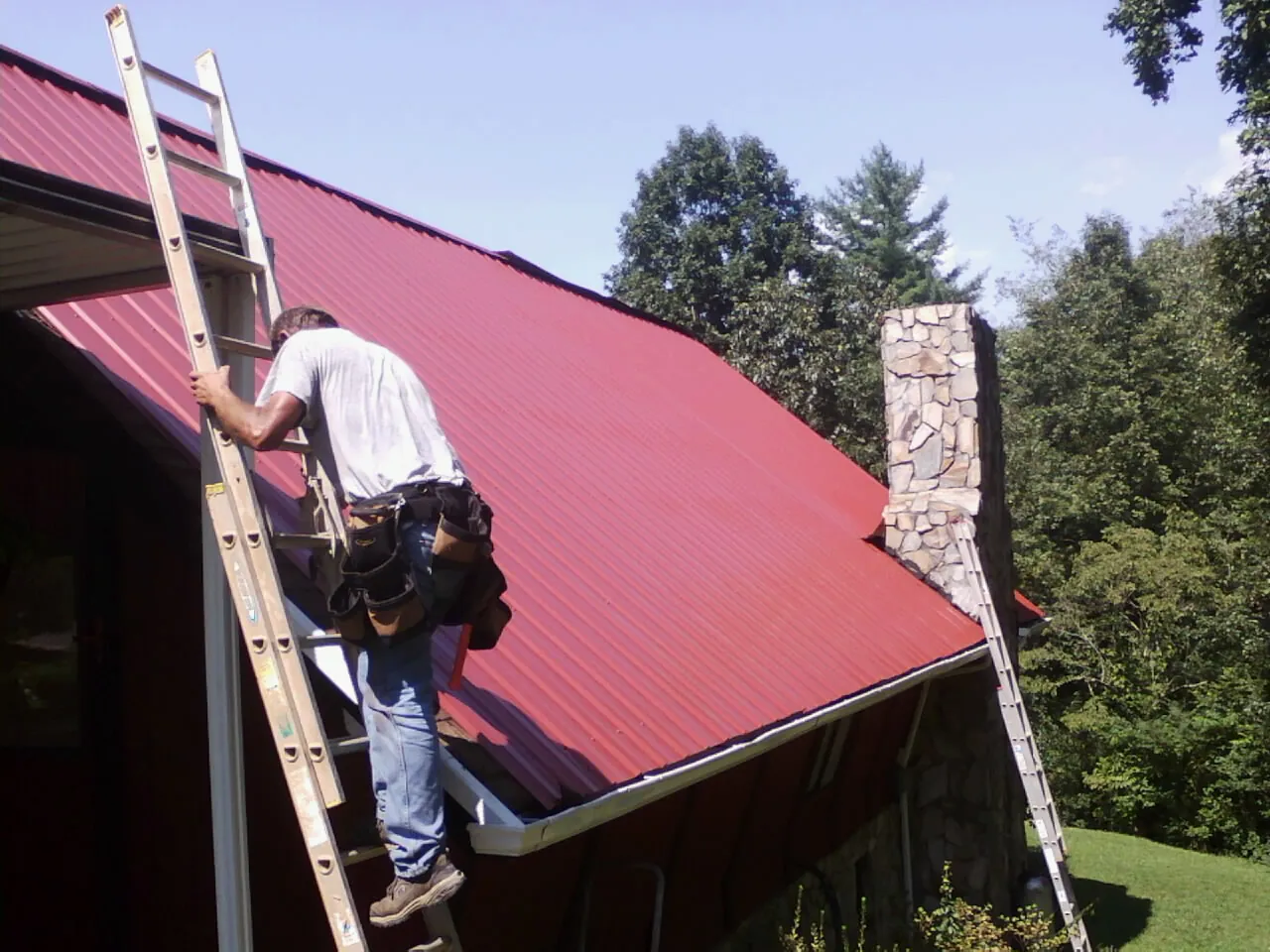 Roofer on a ladder beside a red metal roof and stone chimney — Soffit & Fascia Repair by Roofing Brothers in Amma